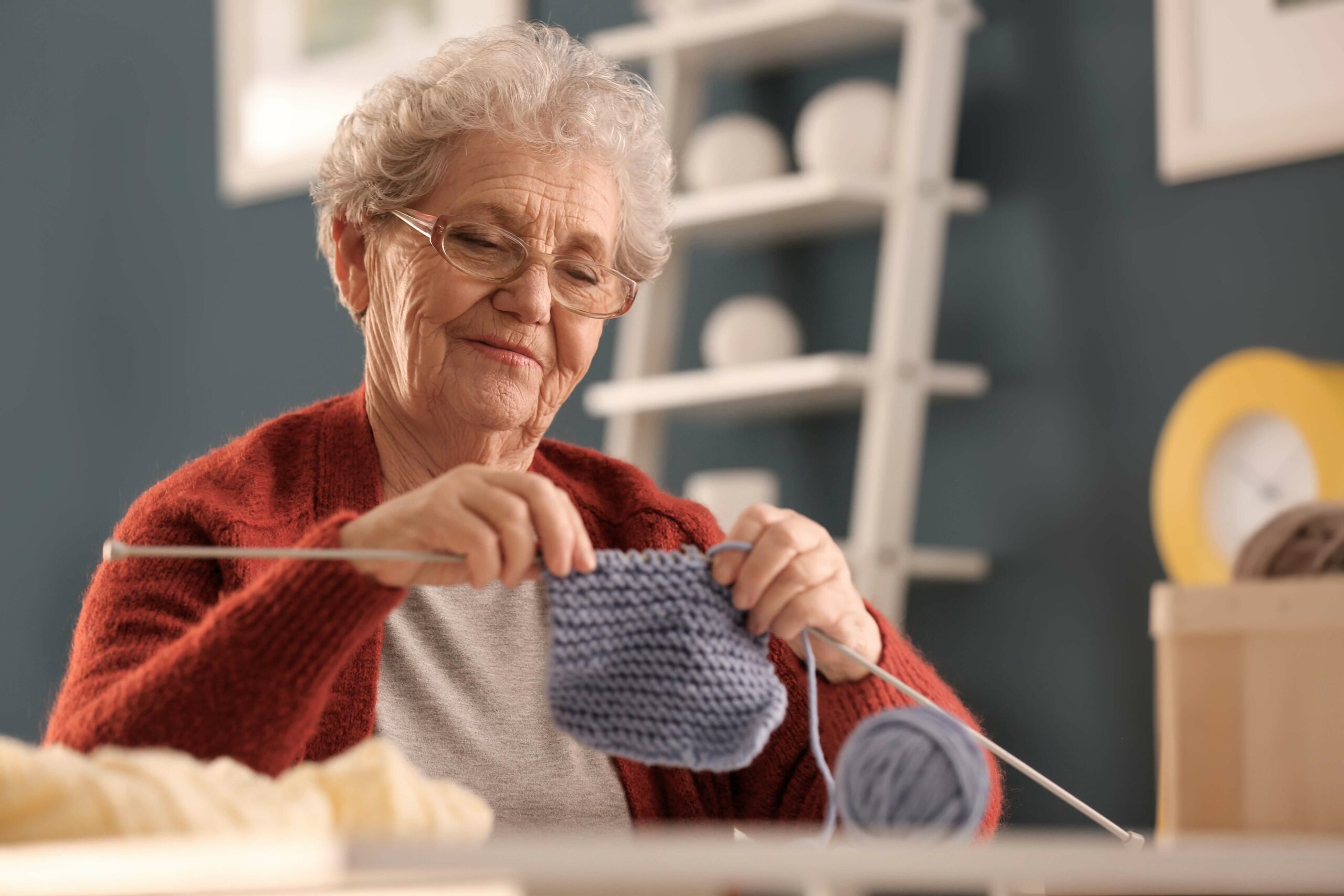 Senior woman knitting with blue yarn, wearing glasses and a red sweater, enjoying a relaxing craft activity in a cozy indoor setting.