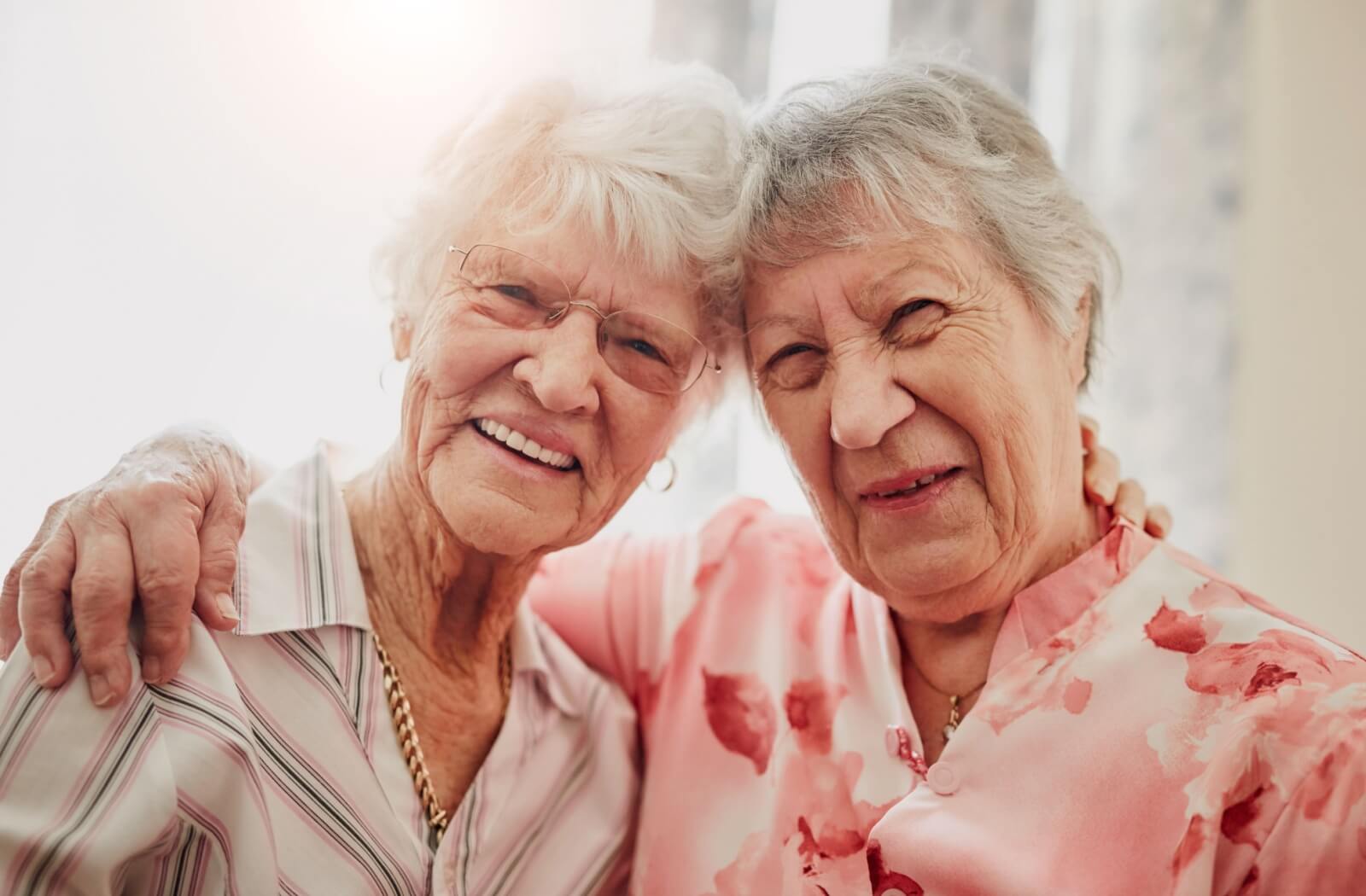 Two senior friends smiling warmly with their arms around each other's shoulders, sharing a moment of friendship