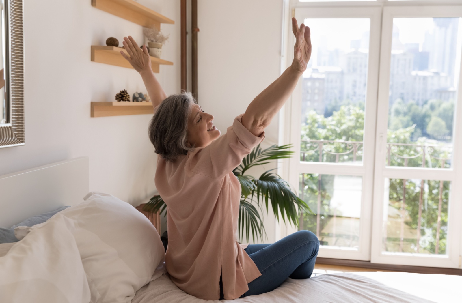 A smiling senior sits on the edge of their bed and stretches in the morning, happy to have so much energy
